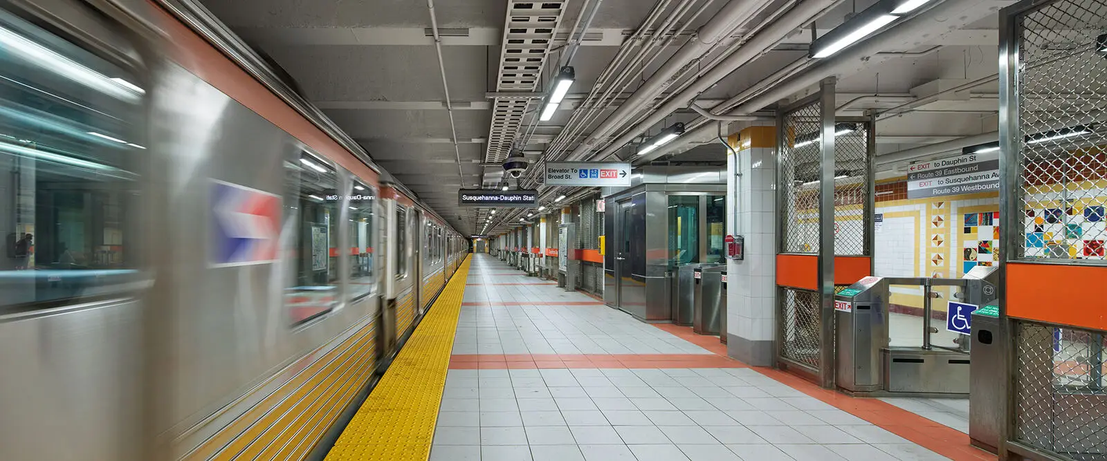 A subway train stops at a clean, modern underground station in Philadelphia with yellow tactile paving, metal fencing, and directional signs. The empty, well-lit platform features accessible entry gates designed by a visionary architect.