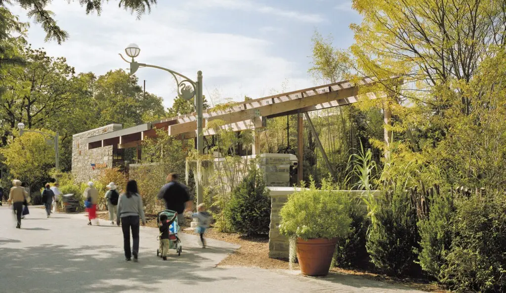 People walk along a paved path lined with greenery and large potted plants, some pushing strollers, near a stone and wood pergola structure designed by an architect committed to historic preservation on a sunny day in Philadelphia.