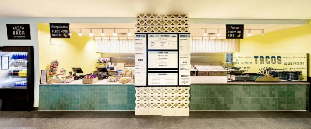 A modern cafeteria counter in Philadelphia features a central menu board, turquoise tiles, snack baskets, a drink cooler on the left, and a food pickup area on the right with TACOS painted on the wall—a showcase of sleek interior design.