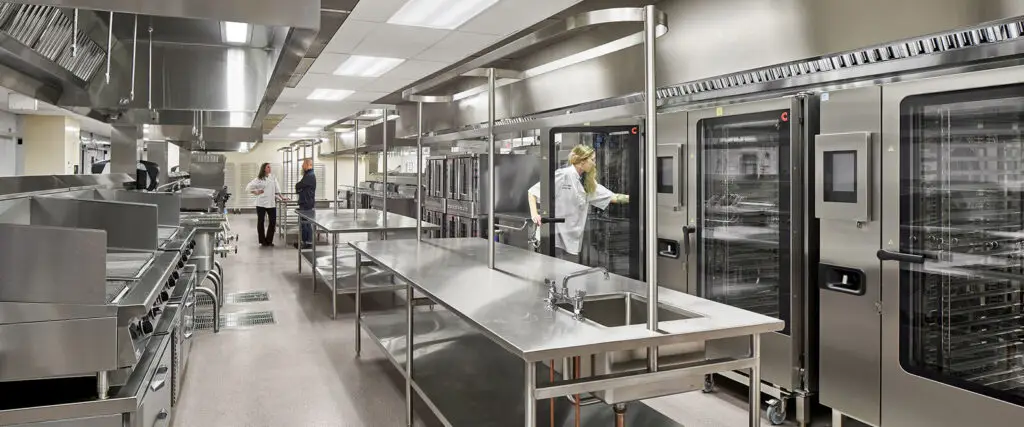 A modern commercial kitchen in Philadelphia features stainless steel work surfaces, large ovens, and industrial appliances. Several people in white coats are working in the clean, brightly lit space with sleek interior design elements.