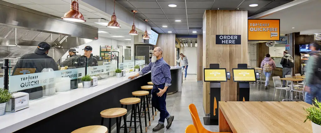 A casual restaurant with an open kitchen, architect-inspired modern decor featuring wood and metal accents, digital order kiosks, customers ordering and waiting, and staff behind the counter.