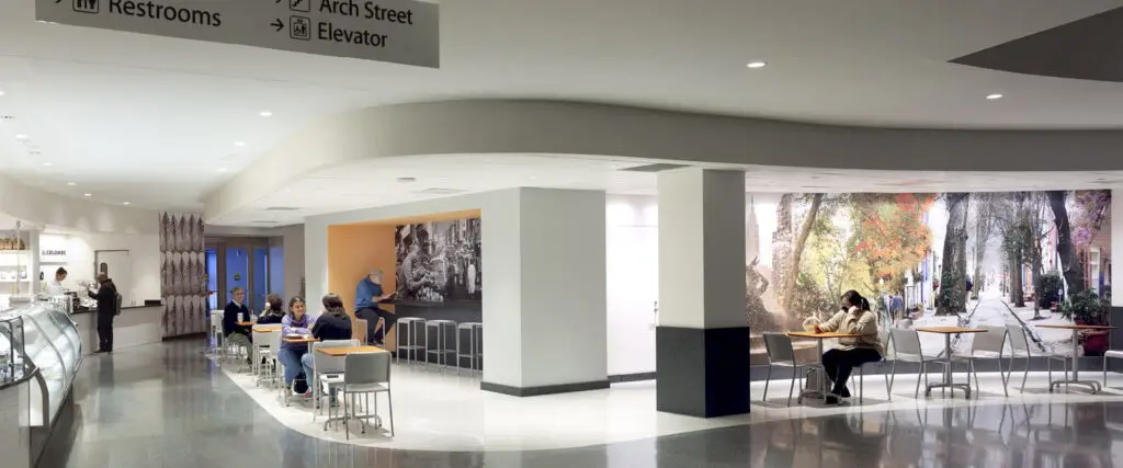 A modern indoor cafeteria in Philadelphia with people sitting at tables and a counter. A mural of a scenic street covers one wall, and signs above direct to restrooms, Arch Street, and an elevator. The civic space is bright and clean.