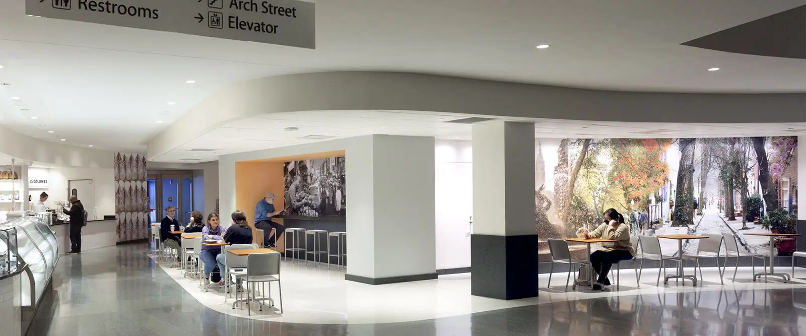 A modern indoor cafeteria in Philadelphia with people sitting at tables and a counter. A mural of a scenic street covers one wall, and signs above direct to restrooms, Arch Street, and an elevator. The civic space is bright and clean.