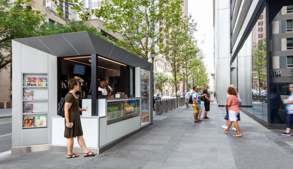 A woman stands at a modern newsstand kiosk on a Philadelphia city sidewalk. People stroll among tall buildings and trees on a sunny day, enjoying magazines and snacks—an educational pause in an urban landscape rich with historic preservation.