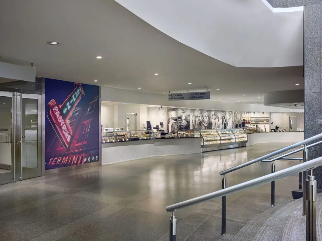 A modern indoor food counter in Philadelphia features glass display cases with pastries and a large mural of neon signs and black-and-white historic photos, blending chic interior design with hints of historic preservation. The area is clean, spacious, and well-lit.