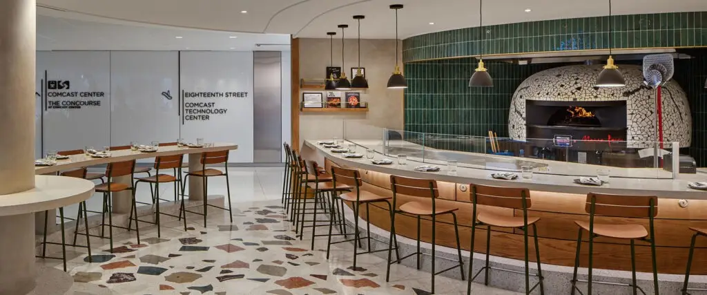 A modern restaurant interior that highlights thoughtful interior design, featuring a curved wooden bar with stools, set tables, a tiled pizza oven with fire burning, and green tiled wall accents. Shelves with books and decor add an educational touch.