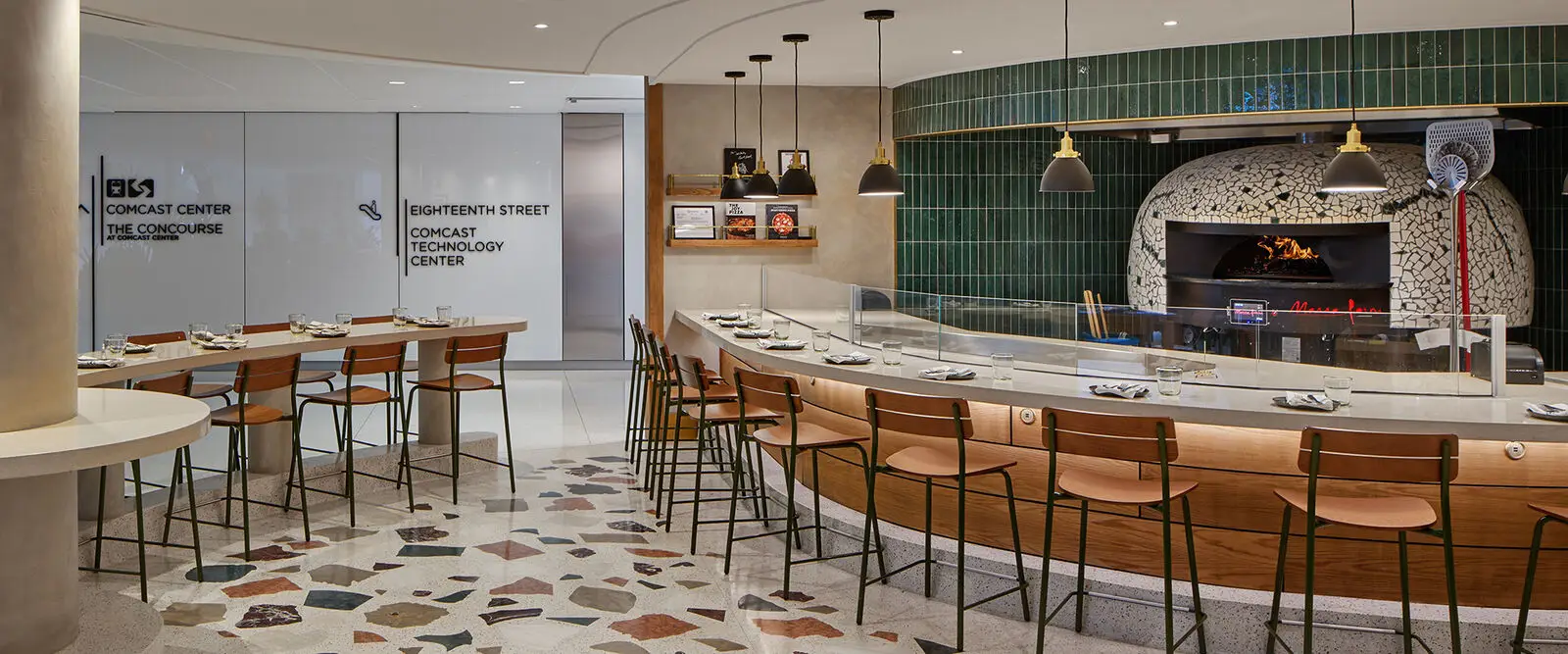 A modern restaurant interior that highlights thoughtful interior design, featuring a curved wooden bar with stools, set tables, a tiled pizza oven with fire burning, and green tiled wall accents. Shelves with books and decor add an educational touch.