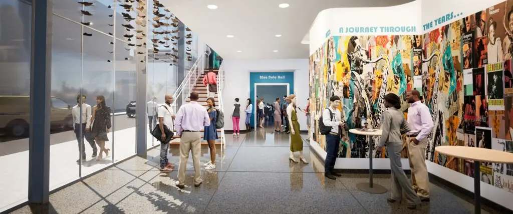 A group of people walk and converse in a bright hallway with floor-to-ceiling windows, modern interior design, and a colorful mural reading “A Journey Through...” on the wall. The lively, educational setting includes some standing by high tables.