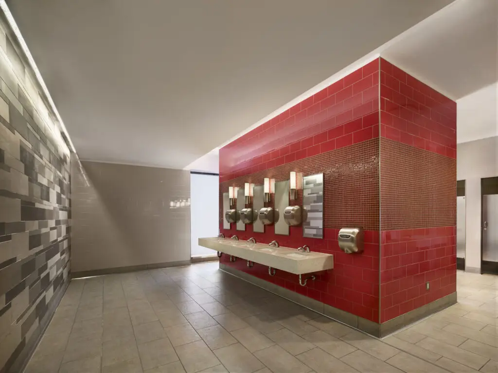 A modern civic public restroom with beige tiled floors, a wall of red tiles surrounding sinks, soap dispensers, and hand dryers, designed with an architect's attention to historic preservation in the beige and brown tiled walls in the background.