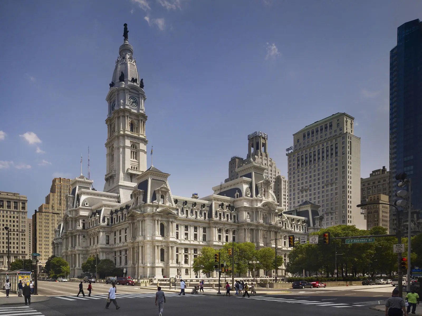 Philadelphia City Hall, designed by a renowned architect, features ornate architecture and a tall clock tower at a busy intersection. Pedestrians cross the street as modern buildings surround this educational city landmark under a clear sky.