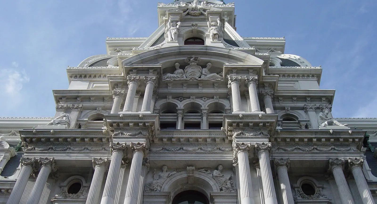 A close-up upward view of a grand, historic building facade in Philadelphia showcases the work of a master architect—ornate columns, arches, carvings, and statues set against a clear blue sky create an awe-inspiring educational sight.