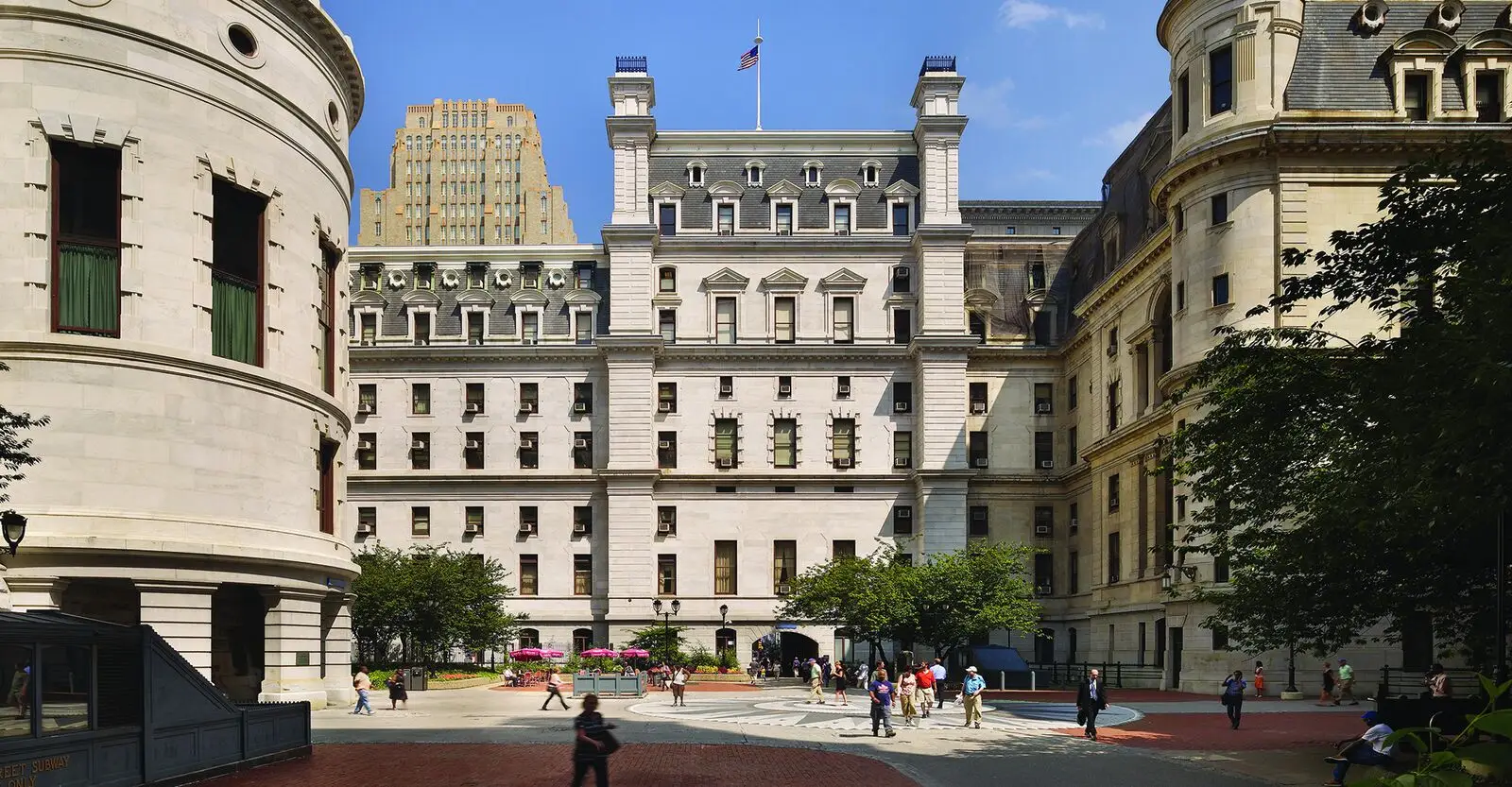People walk through a sunny courtyard surrounded by historic stone buildings, celebrated for their architect's skill in historic preservation; arched windows and columns frame the scene as a U.S. flag flies above and trees and benches invite visitors to linger.