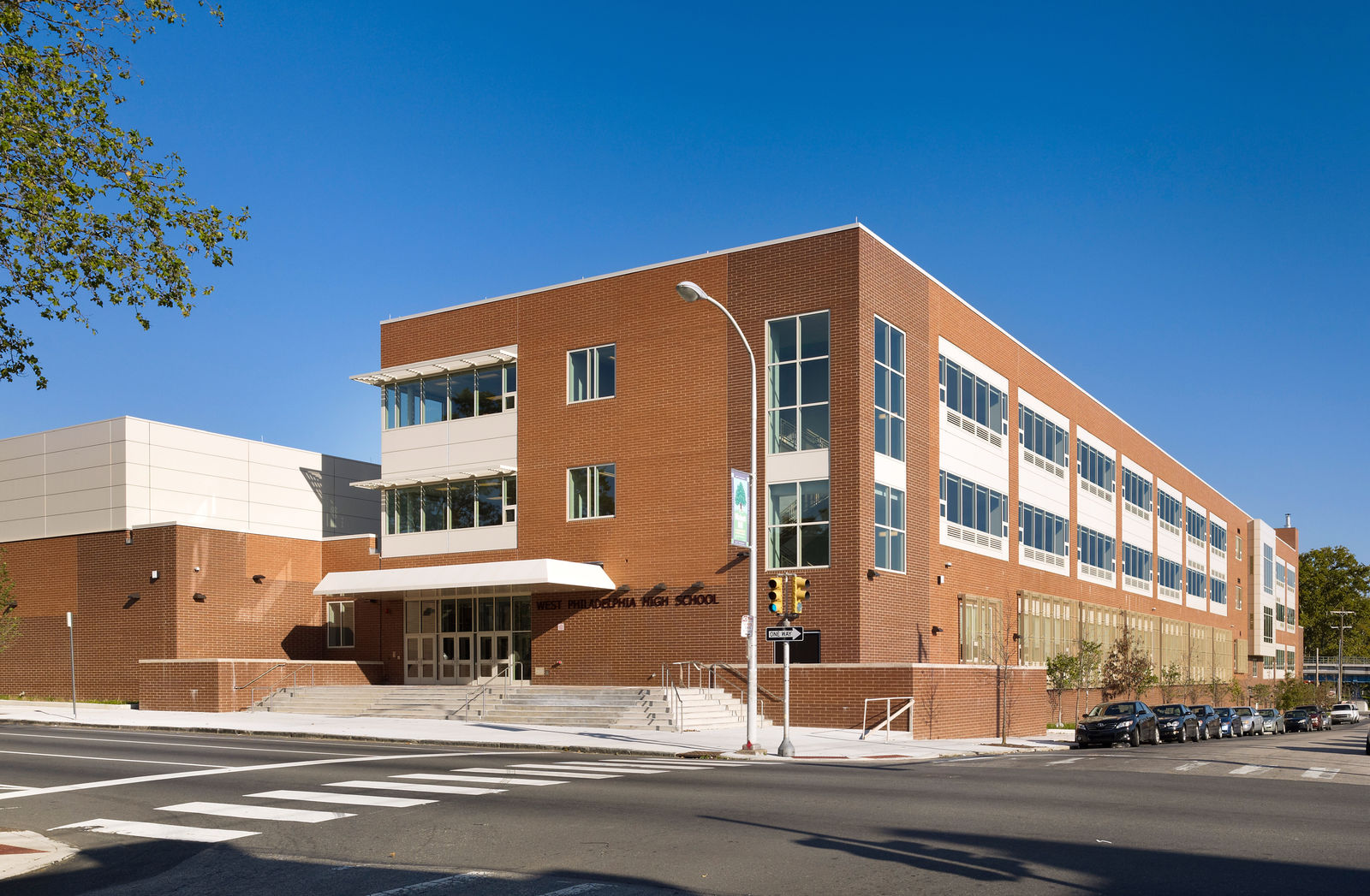 A modern three-story brick school building with large windows, designed by a noted architect, sits on a Philadelphia street corner under a clear blue sky. Cars are parked alongside and a crosswalk leads to this civic landmark’s entrance.