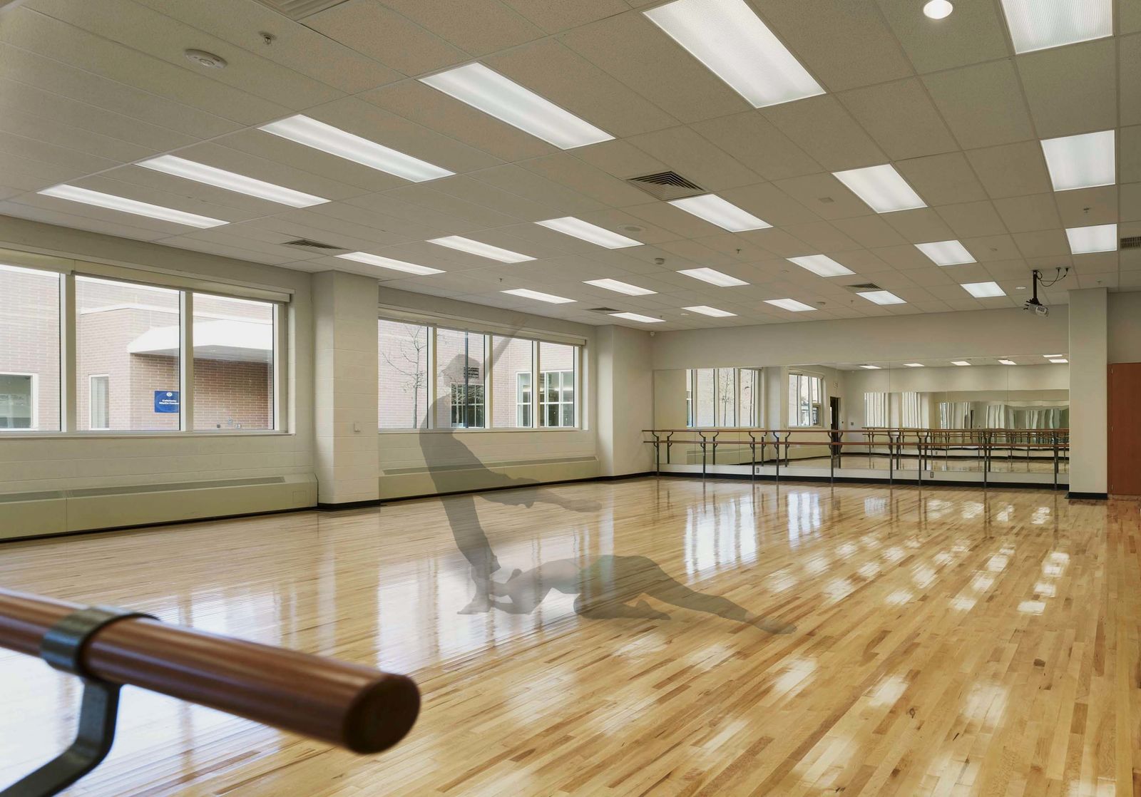 A bright, spacious dance studio with large windows, wooden floors, mirrors on one wall, and ballet barres showcases thoughtful interior design. A person is performing a breakdancing move in the center of the room.