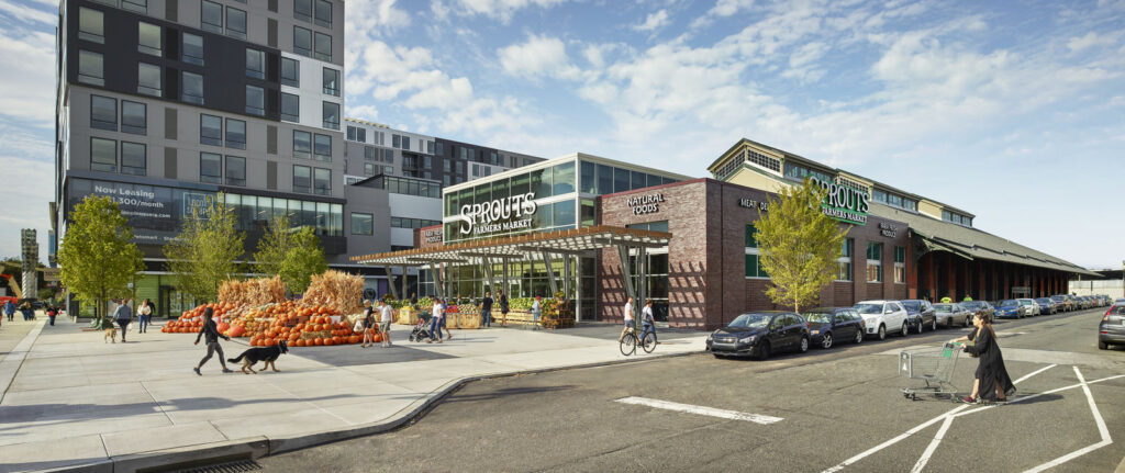 A modern city street with a Sprouts Farmers Market grocery store, outdoor produce display with pumpkins, pedestrians, cyclists, parked cars, and a person pushing a shopping cart under a partly cloudy sky in a lively civic setting.