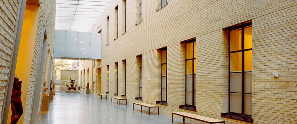 A spacious, modern hallway in Philadelphia with tall windows, beige brick walls, a glass ceiling, and benches along the wall. Sculptures are visible at the far end and left side, with natural light emphasizing thoughtful interior design.
