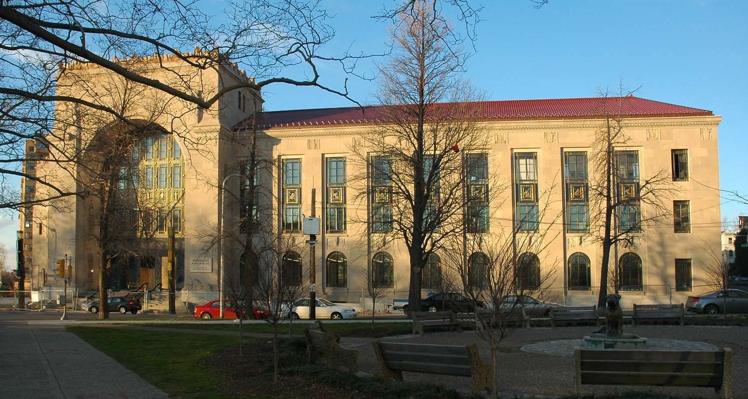 A large, beige stone building with tall arched windows, a red-tiled roof, and decorative columns reflects Philadelphia's civic pride and commitment to historic preservation. Trees, benches, and several parked cars line the sunny street in front.