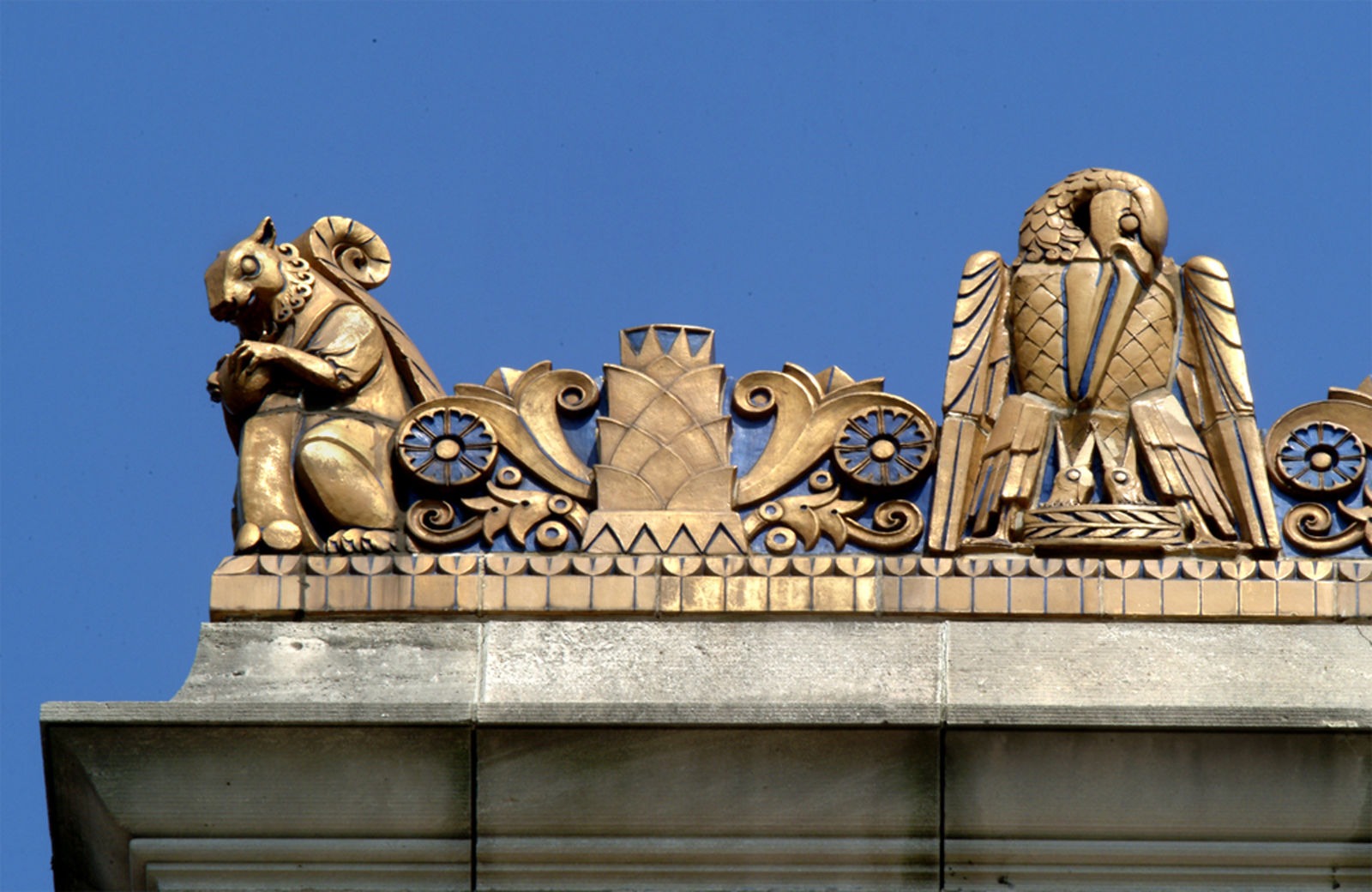 Gold ornamental rooftop sculptures against a blue sky, designed by a renowned architect, feature a stylized squirrel holding an acorn on the left and an eagle on the right, separated by intricate decorative scrollwork.