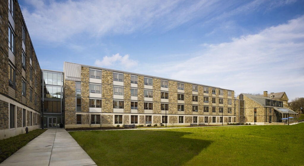 A modern, four-story building designed by a renowned architect features a stone and glass exterior, nestled beside a manicured lawn in Philadelphia. A sidewalk leads to the entrance on the left side under blue skies with scattered clouds.