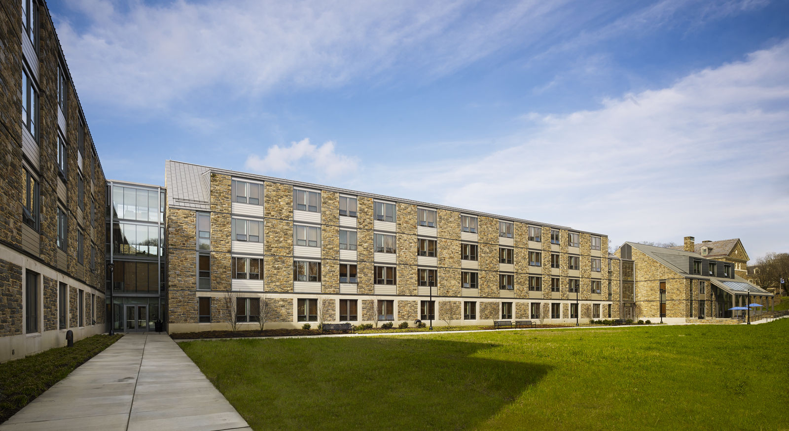 A modern, four-story building designed by a renowned architect features a stone and glass exterior, nestled beside a manicured lawn in Philadelphia. A sidewalk leads to the entrance on the left side under blue skies with scattered clouds.