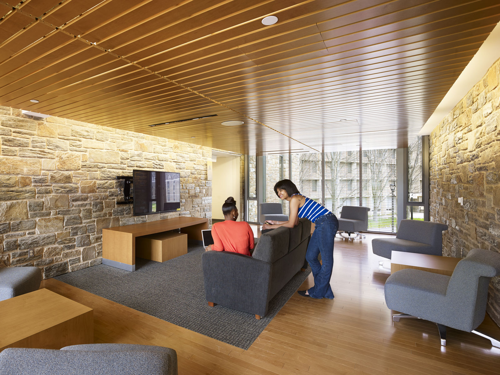 Two people work on a laptop in a modern lounge in Philadelphia, where thoughtful interior design blends stone walls, large windows, and gray furniture. Natural light fills the space, creating a bright and welcoming atmosphere.