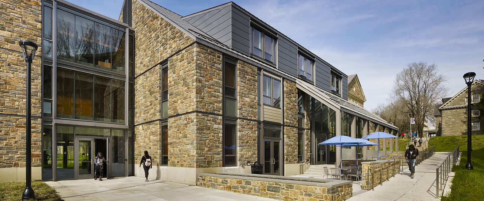 A modern educational building with large windows and stone walls, designed by a renowned architect in Philadelphia, features outdoor seating with blue umbrellas as people stroll along the sidewalk under a clear, sunny sky.