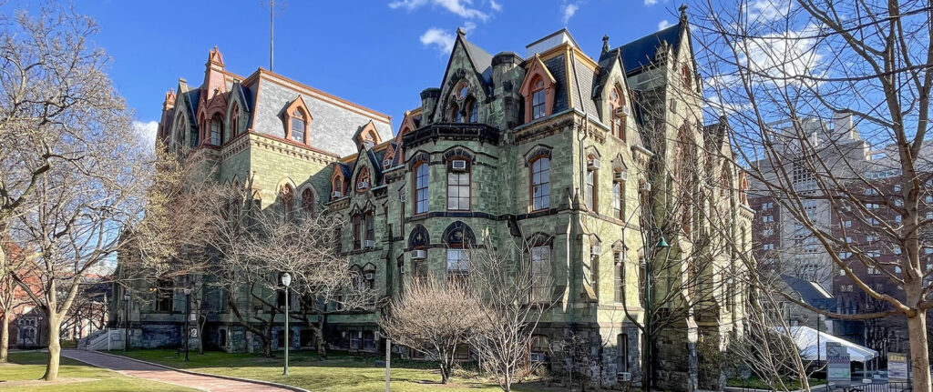 A large, historic stone building with ornate architectural details, arched windows, and pointed roofs showcases the work of a skilled architect, surrounded by bare trees and a brick pathway under a blue sky with scattered clouds.