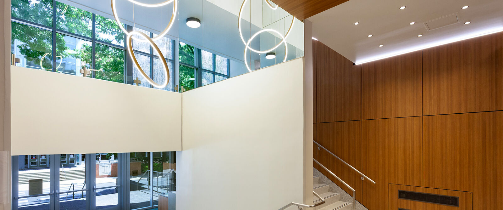 A modern building interior with large windows, wooden wall panels, circular pendant lights, and a staircase leading to an upper floor. Sunlight streams through the windows, highlighting thoughtful interior design details throughout the space.