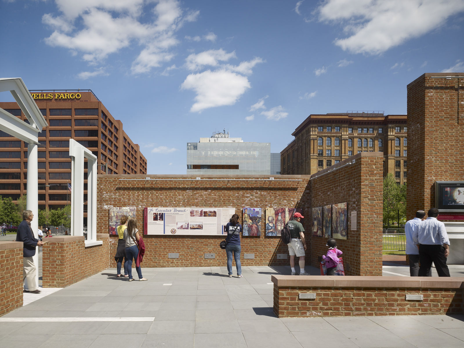 People view historical displays on brick walls in an open-air plaza, surrounded by modern buildings under a blue sky. In this civic space in Philadelphia, the Wells Fargo building stands tall, highlighting the city’s commitment to historic preservation.