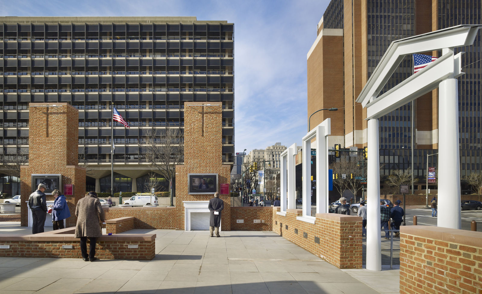 People explore an outdoor historical site with brick walls, columns, and American flags. This civic space highlights historic preservation amid modern office buildings and traffic under a blue sky.