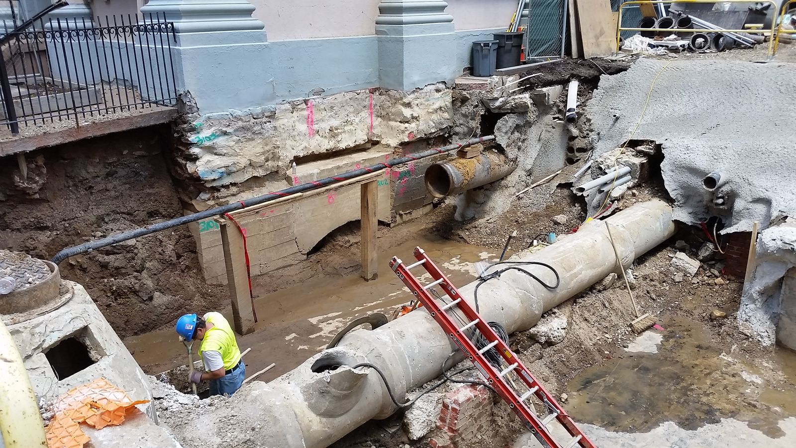 A construction worker in a yellow vest and blue helmet stands in a large muddy excavation site with exposed pipes, concrete walls, and ladders near a building’s foundation, aiding civic development in Philadelphia.