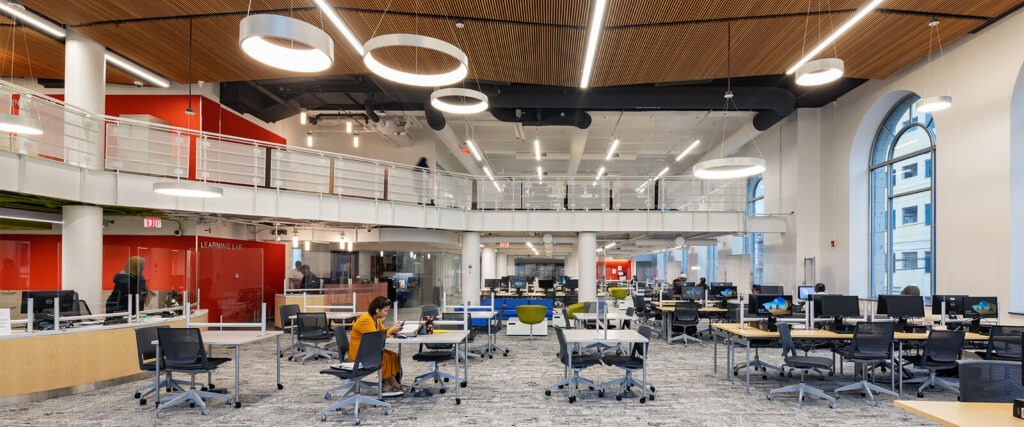 A modern, open-plan office designed by an architect features rows of desks, computers, and office chairs. One person sits working while others move about. Large windows and a mezzanine level create a bright, educational atmosphere.