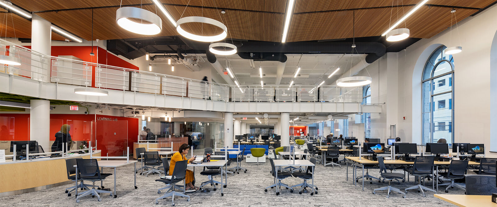 A modern, open-plan office designed by an architect features rows of desks, computers, and office chairs. One person sits working while others move about. Large windows and a mezzanine level create a bright, educational atmosphere.