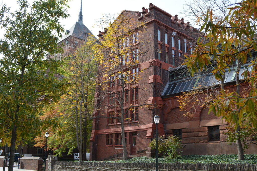 Red brick academic building partially covered by autumn trees, with large windows and a spire of another educational building in the background. A black iron fence and streetlamp are in the foreground.