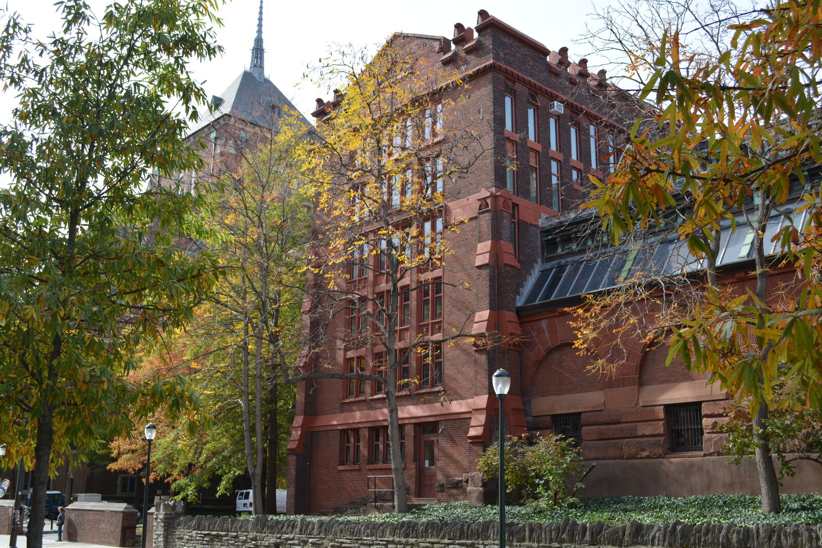 Red brick academic building partially covered by autumn trees, with large windows and a spire of another educational building in the background. A black iron fence and streetlamp are in the foreground.