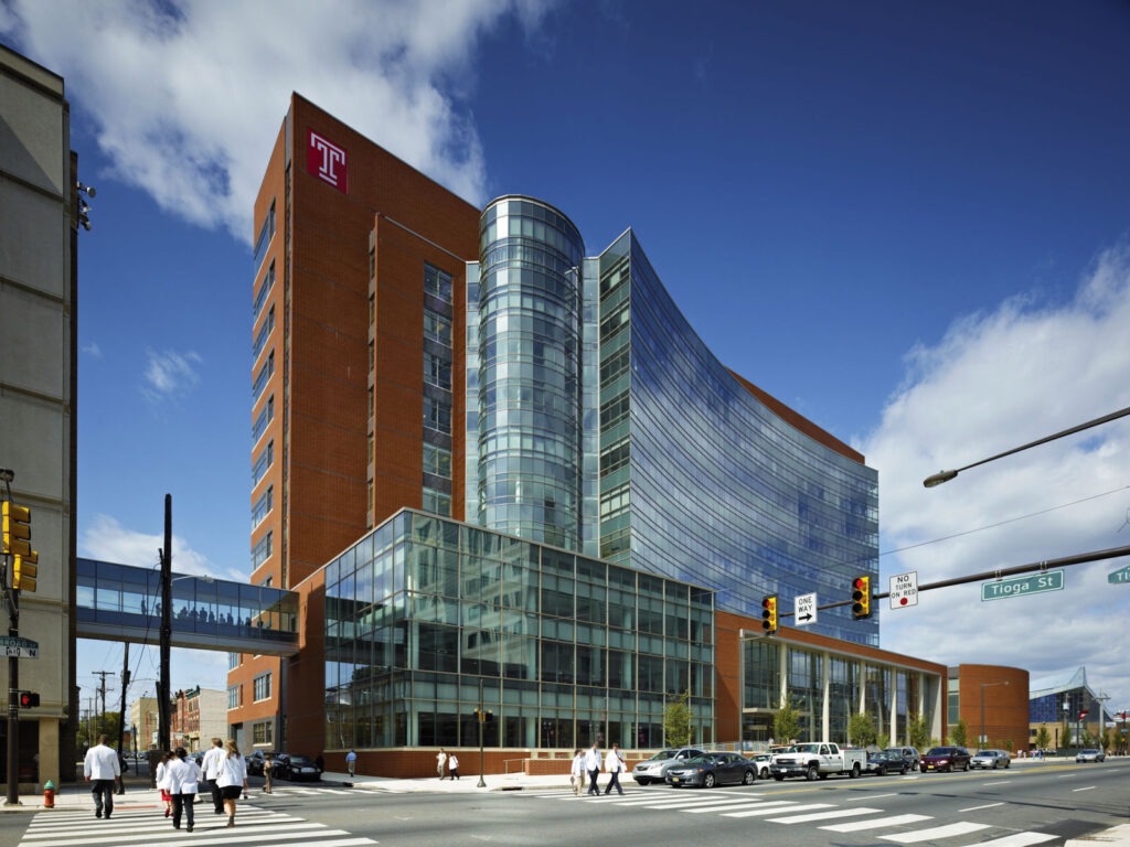 A modern, multi-story glass and brick building designed by an architect, featuring a Temple University T logo at a busy Philadelphia intersection, with a skywalk connecting to another educational building and people crossing the street.