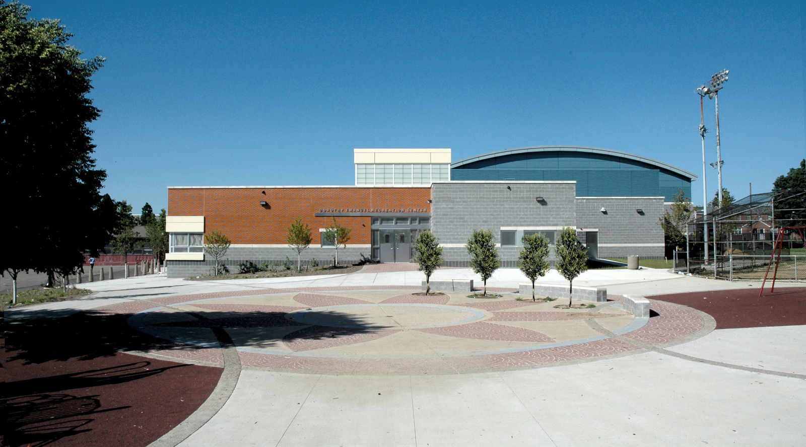 A modern civic community center building in Philadelphia with gray, blue, and orange panels, large windows, and a circular patterned plaza surrounded by small trees, showcasing thoughtful interior design under a clear blue sky.