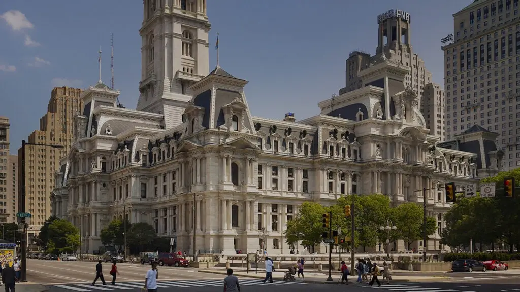 A historic, ornate stone building with a tall clock tower, designed by a renowned architect, stands at a busy civic intersection, surrounded by modern skyscrapers and people crossing the street in daylight.