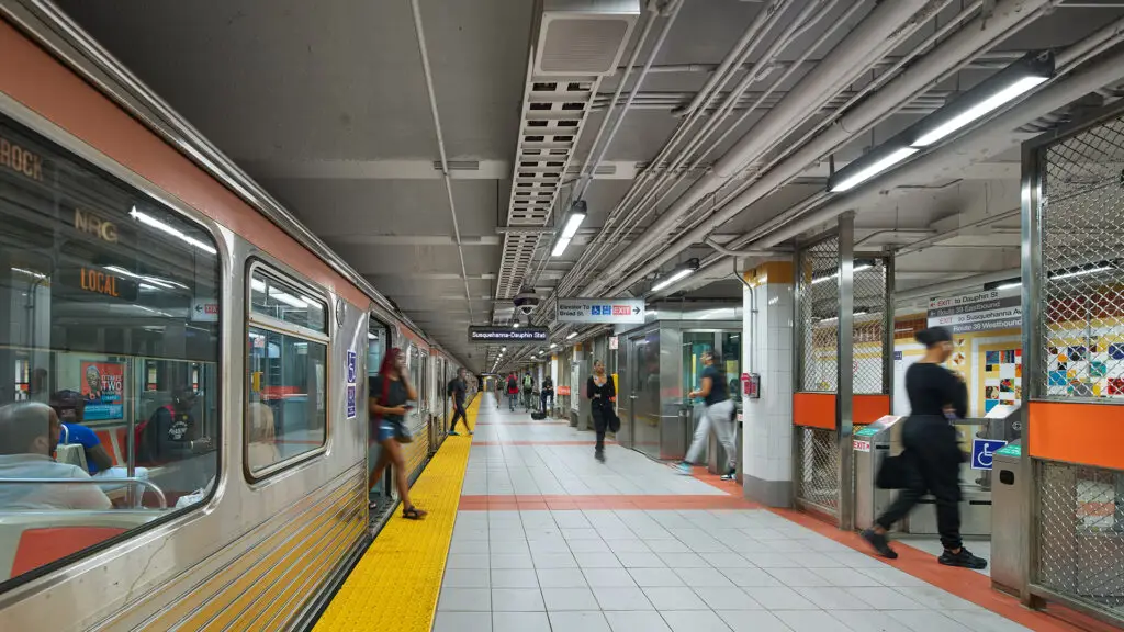 Passengers wait and walk along a well-lit Philadelphia subway platform, where colorful tiles and overhead pipes reflect thoughtful interior design. A train is stopped on the left side, with signs and station details framing the scene.