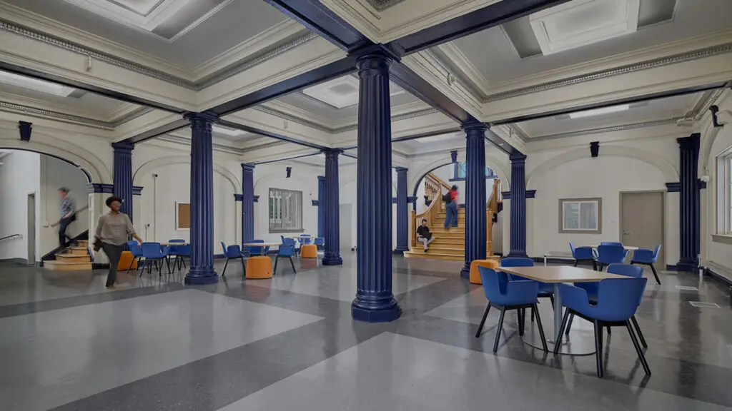 A spacious, modern civic interior in Philadelphia with tall blue columns, round tables with blue chairs, grey floors, and people moving about. Stairs lead to an upper level where large windows flood the space with natural light.
