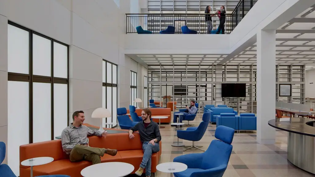 A modern, spacious lounge with large windows, orange and blue seating, white tables, and a mezzanine level where several people are talking or working. This educational space is filled with natural light as people engage in conversation.