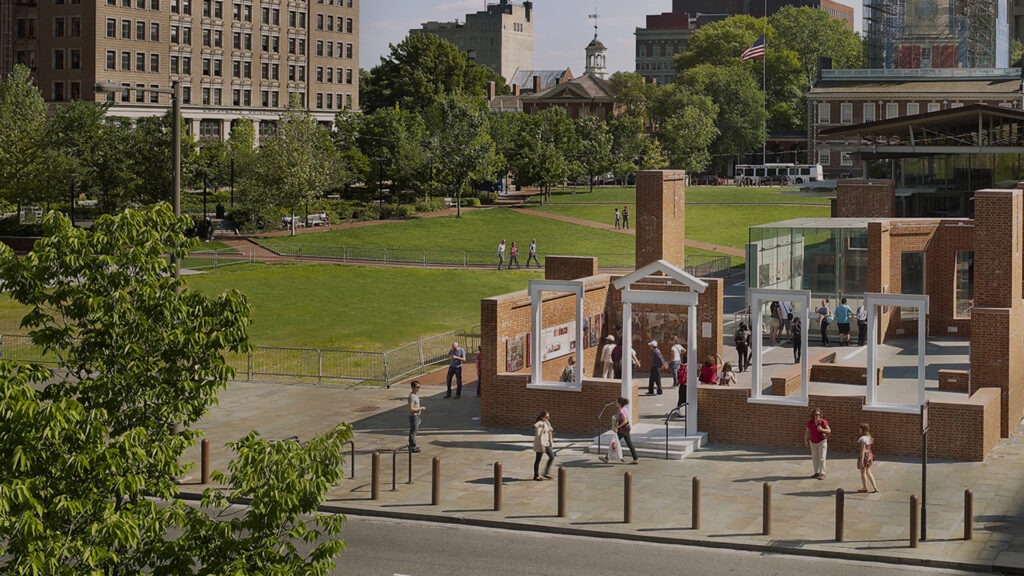 A city park with people walking and relaxing; in the foreground is an open-air brick structure curated for historic preservation, resembling a partial house or historical site, with tall buildings and trees in the background.