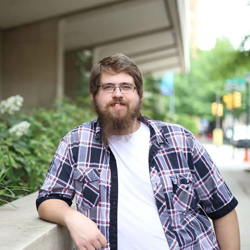 A bearded man wearing glasses and a plaid shirt over a white t-shirt leans against a concrete ledge outdoors in Philadelphia, with greenery and a blurred city street in the background.
