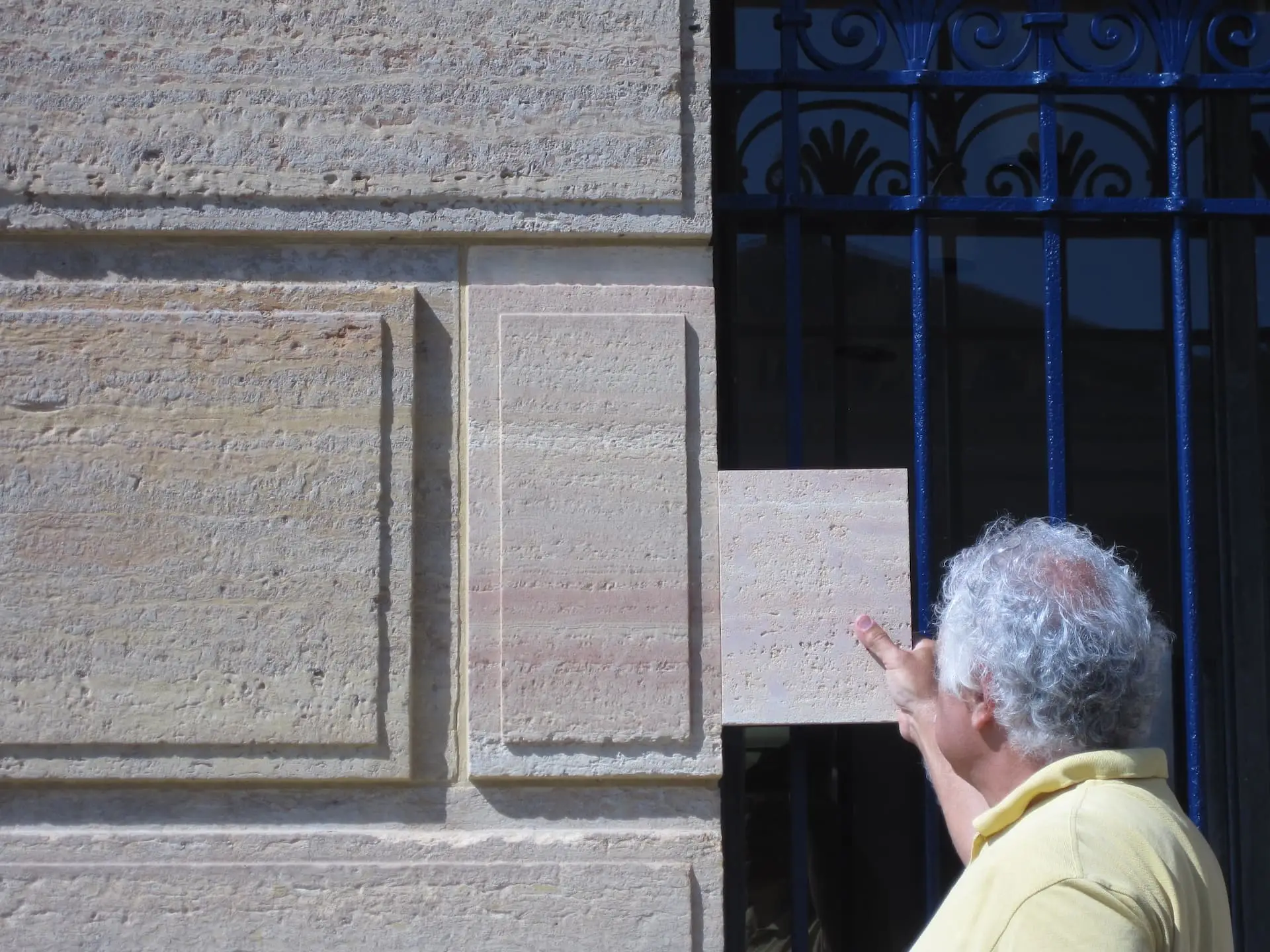 A person with gray hair and a yellow shirt points at a rectangular stone panel partly pulled out from a textured stone wall, near a blue metal fence, highlighting historic preservation in Philadelphia.