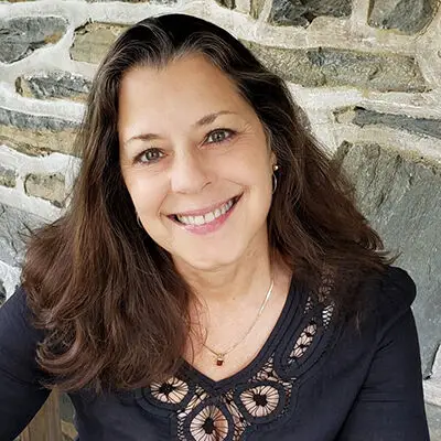 A woman with long brown hair and a black top smiles while sitting in front of a stone wall, creating an inviting scene with a touch of interior design flair.