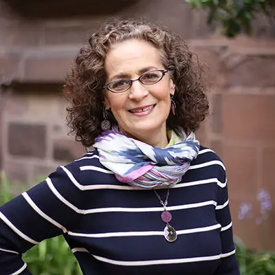 A woman with curly brown hair and glasses smiles while standing outdoors. Showcasing an educational flair, she wears a black and white striped top, colorful scarf, pendant necklace, and earrings. A blurred brick wall and greenery are in the background.