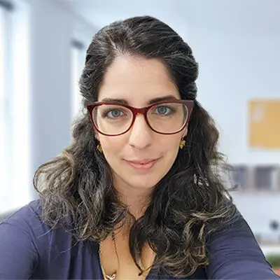 A woman with long, wavy dark hair and red-framed glasses smiles slightly in an educational office setting in Philadelphia, with bright windows and a corkboard in the background.
