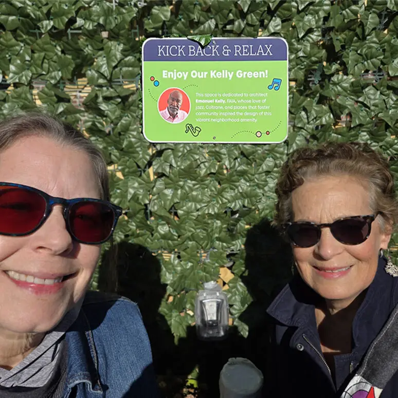 Two women wearing sunglasses smile for a selfie in front of a leafy green wall with a sign reading “KICK BACK & RELAX. Enjoy Our Kelly Green!” featuring civic information about architect Kelly.