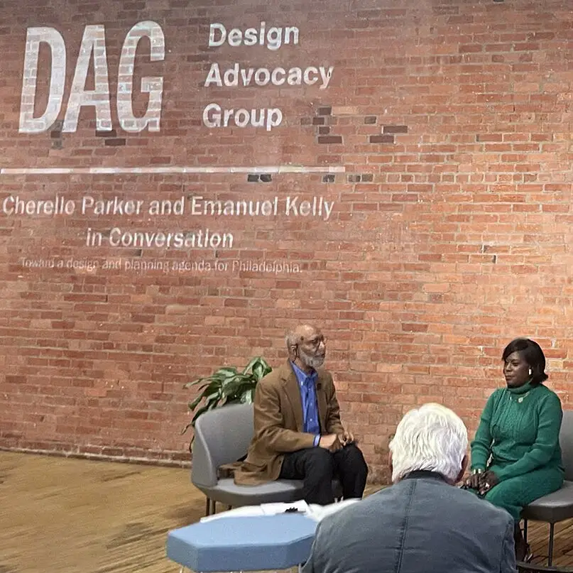 Two people sit facing each other onstage in front of a brick wall with “Design Advocacy Group” and event details displayed, engaging in an educational discussion about historic preservation in Philadelphia as an audience member listens intently.
