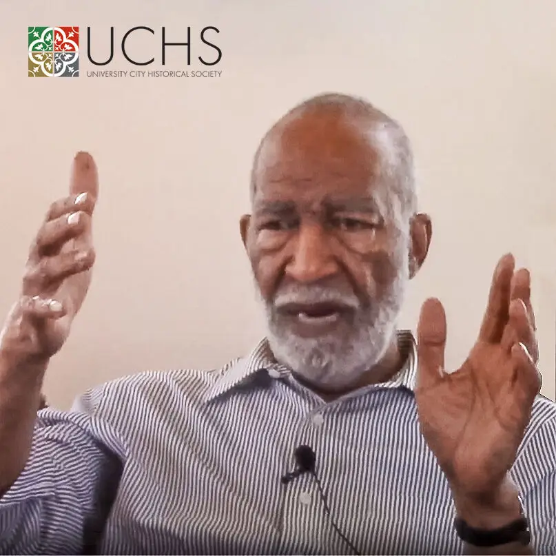 An older man with a gray beard gestures with both hands while speaking about historic preservation. He wears a striped shirt and has a microphone clipped to his collar. The UCHS (University City Historical Society) logo is in the top left corner.